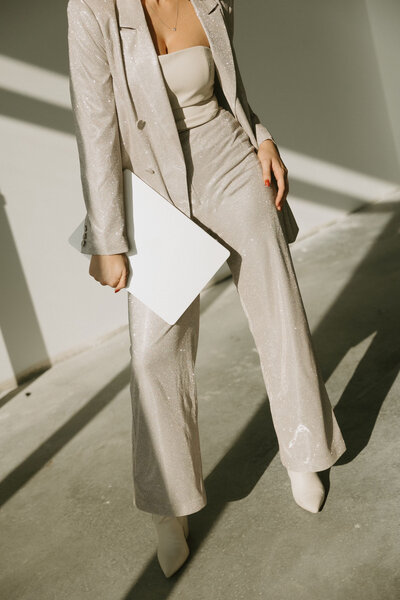 Professional woman in beige blazer and matching pants holding white folder, photographed in natural light with architectural shadows.