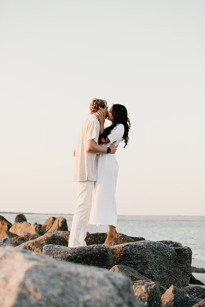 Dark haired women in white dress and man in white pants and beige shirt kissing on rocks