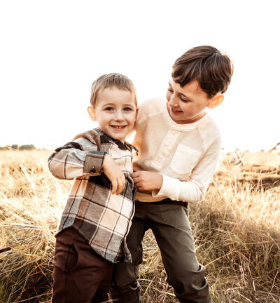 A photo of two boys smiling one waring brown flannel and brown jeans and the other wearing tan shirt and green pants
