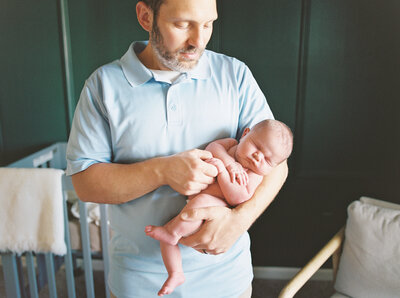 Father in baby blue shirt holds naked newborn son while looking at him adoringly
