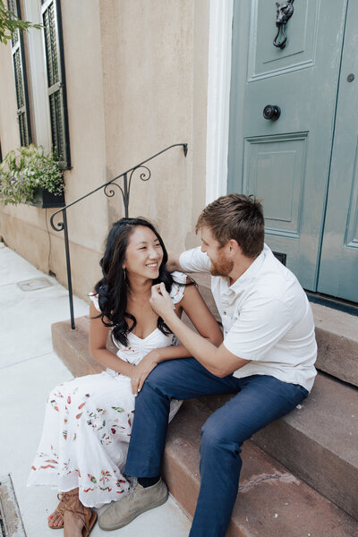 Couple sitting on steps wearing white and jeans laughing together in historic downtown Charleston during engagement photos.