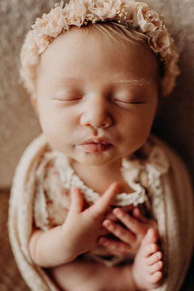 A perfectly posed newborn, baby girl, puckers her lips, while she is in an open swaddle, wearing a peach colored floral crown posing perfectly with her eyes closed.