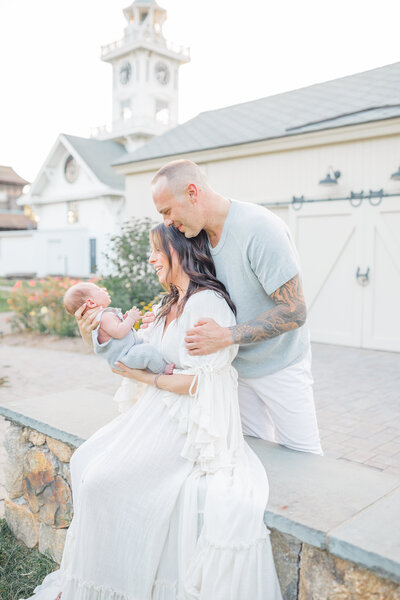 Mom sitting on a patio, gently holding her swaddled newborn in her arms, gazing down at him with a loving expression. Dad stands behind her, looking over her shoulder with a proud and tender gaze, creating a warm family moment outdoors
