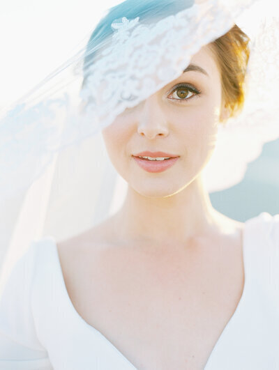 Kohnur bride with natural, soft makeup and elegant updo behind a lace veil, wearing a long-sleeved gown at
