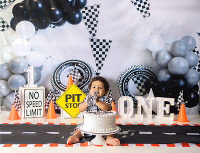 Happy baby boy celebrating his first birthday in race car themed bath setup with arms raised after cake smash in Waldwick NJ studio