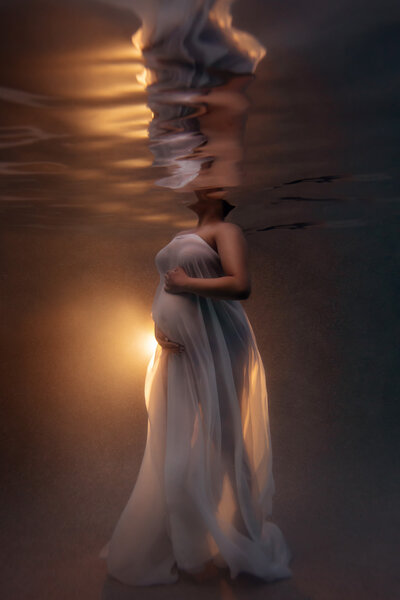 Underwater pregnancy photo with woman draped in white sheer fabric with a light coming from behind and her silhouette reflected on the water taken by Lindsay Walden Photography