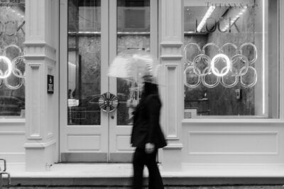 black and white image of woman walking in the rain in front of a storefront