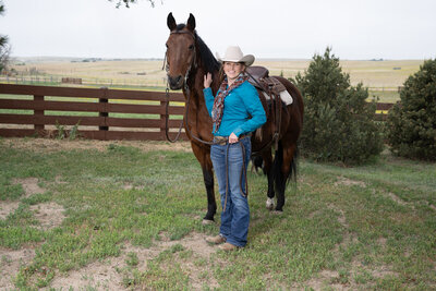 A woman stands with hand on horses neck while she smiles at the camera in western attire