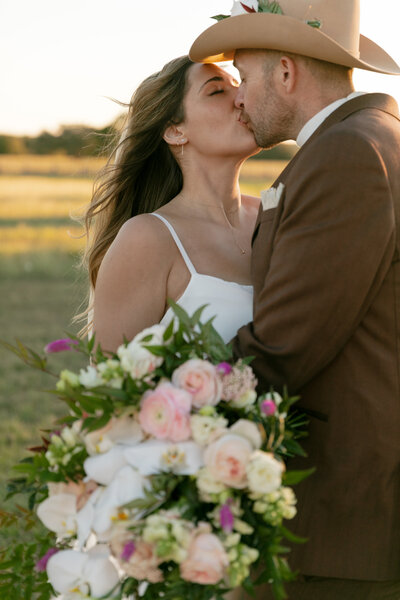 Bride holding her bouquet and groom in his cowboy hat kissing each other. 