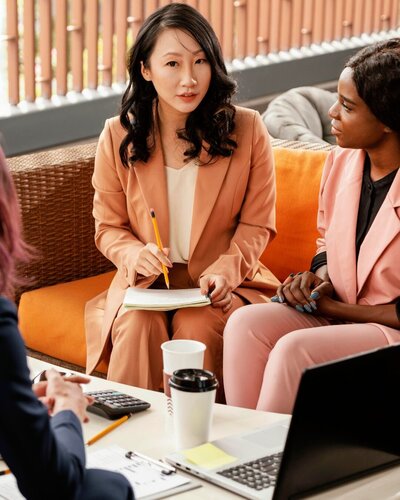 TWO WOMEN IN COLOURED SUITS SAT DOWN CHATTING WITH NOTEBOOK AND PEN