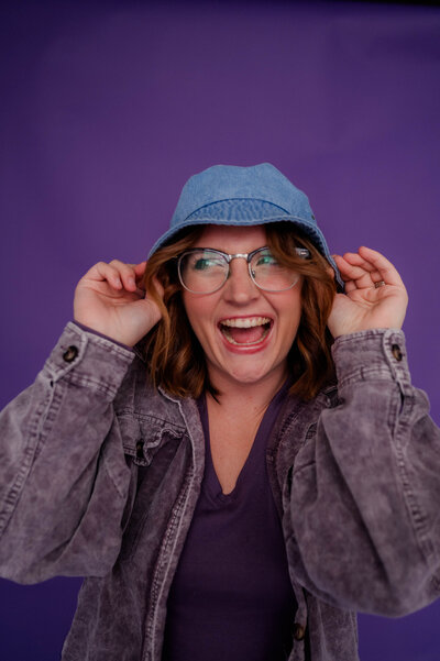 Woman poses for the camera with her hand in her hand wearing denim in front of a purple backdrop.