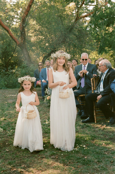 Two young flower girls in white dresses walk down a forest aisle during a Seattle wedding ceremony, captured with soft natural light and documentary-style wedding photography.