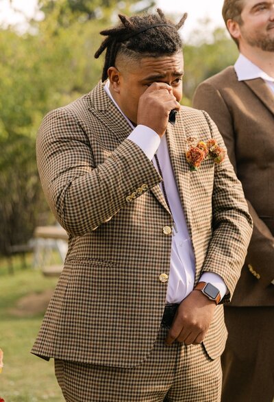 Groom crying happy tears and wiping his eyes when he sees his bride come down the aisle at Venue On The Rocks for their outdoor ceremony