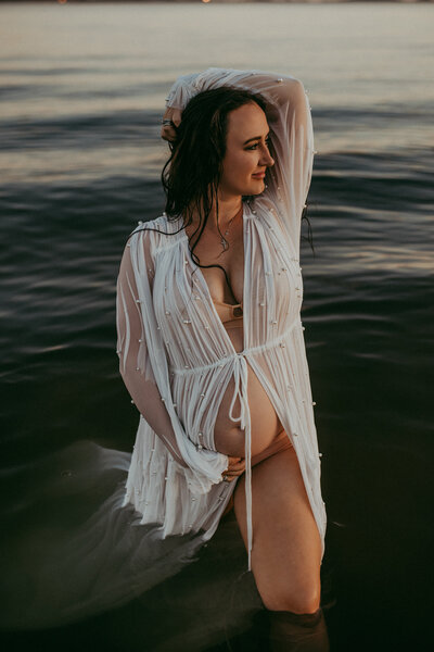 A pregnant mother in a sheer white robe poses in the ocean looking into the sunset while holding her bump. 