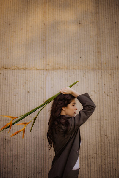 Woman in a brown blazer holds bird of paradise stems over her shoulders, standing against a textured concrete wall.