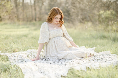 Red-haired pregnant woman in a floral dress sitting on a lace blanket outdoors during a golden-hour maternity session with a Kansas City maternity photographer.