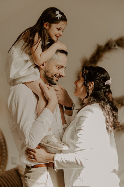 Homme portant sur ses épaules sa fille et regardant sa femme dans un décor intérieur cosy. Photo capturée par Laura, photographe en Vendée.