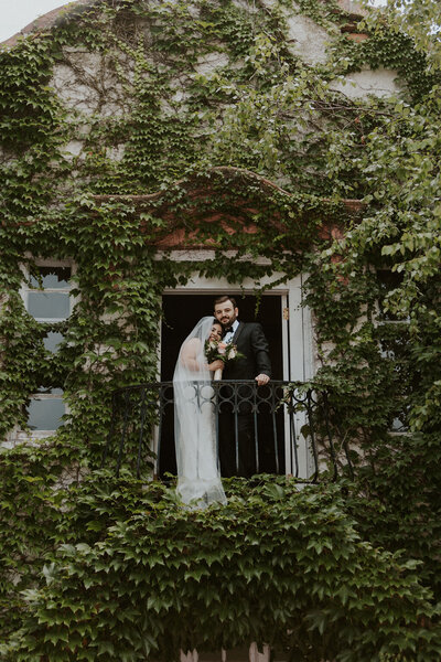 Bride and groom standing on balcony leaning together in vine covered wedding venue
