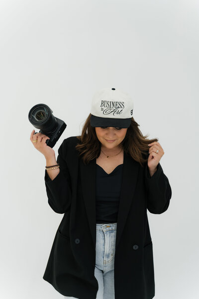 Photographer stands confidently in a white hat that says “Darling Darlene,” holding a camera in one hand against a light gray studio backdrop.
