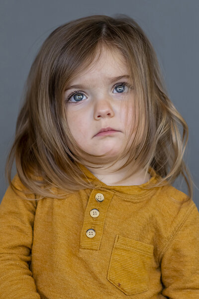 Preschool girl in a mustard yellow top with brown hair, not smiling, but has big blue eyes looking above the camera for her school photo in Port Coquitlam.
