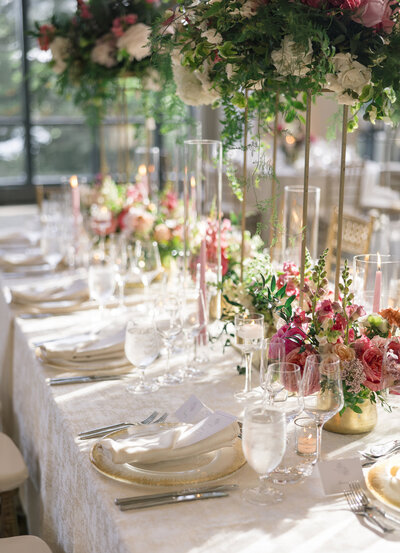 A beautifully set, elegant dining table with white tablecloths is adorned with plates, glasses, and silverware. Tall floral arrangements with greenery and pink flowers rise above, while soft natural light filters through large windows.