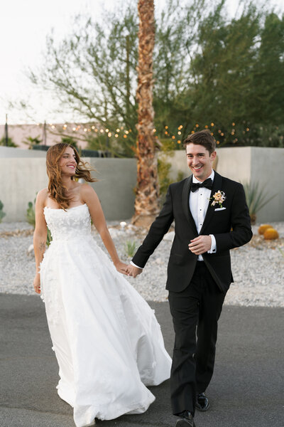 Newlywed couple walking hand-in-hand, smiling joyously, outside of their Palm Springs wedding venue, with the bride's white gown and the groom's black tuxedo against a desert landscape and string lights at dusk.