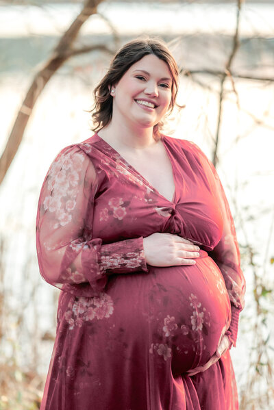 A pregnant woman wears a pink floral dress and smiles for the camera during her Chesapeake, VA maternity session.