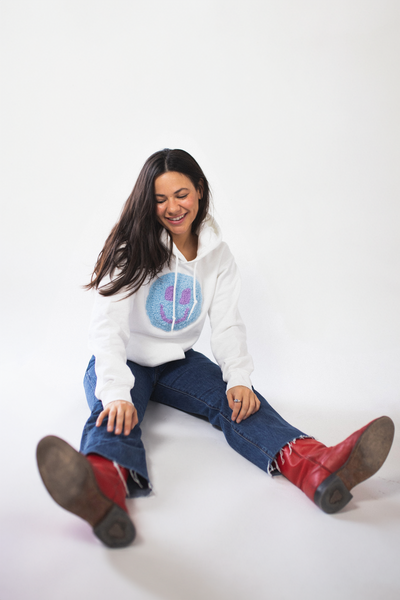 A woman with long dark hair smiles while sitting casually on a white studio floor, wearing a white hoodie with a fuzzy blue and purple smiley face design, red boots, and frayed-hem jeans.