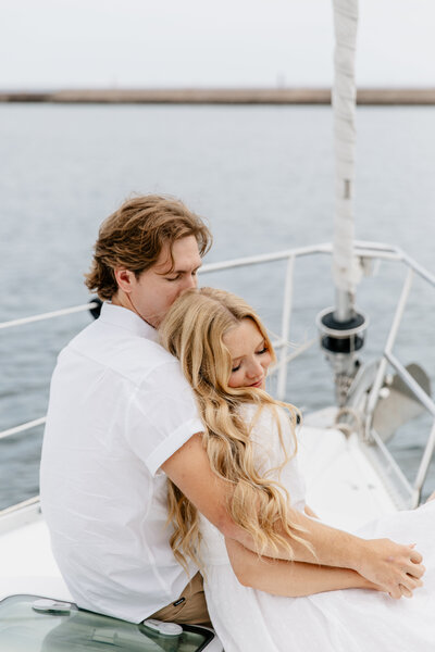 Engaged couple on a sailboat in Grand Marais, MN, celebrating a proposal on Lake Superior, captured by a wedding and elopement photographer in Duluth, MN. Both wearing white, they hold hands as she smiles and he kisses the top of her head, with the serene lake as a backdrop.