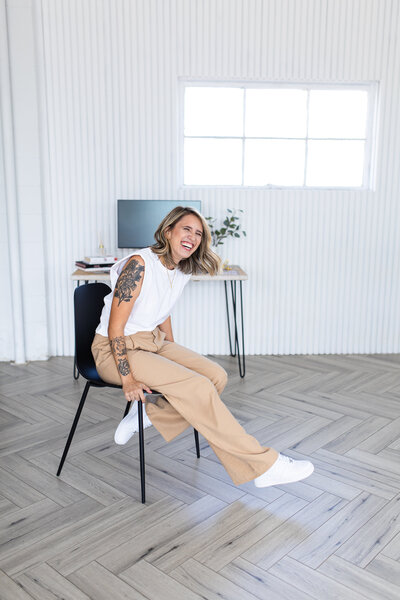 The brand and web designer is sitting on a modern black chair, behind is a wooden furniture with a plasma TV and a white textured wall. She is dressed in beige pants and a white t-shirt.