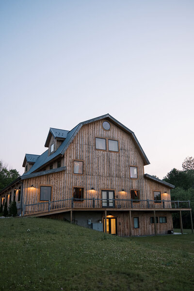 Rustic barn-style venue glowing at dusk with warm lights and wood siding, nestled on a grassy hill at Bear Ridge Destination.

