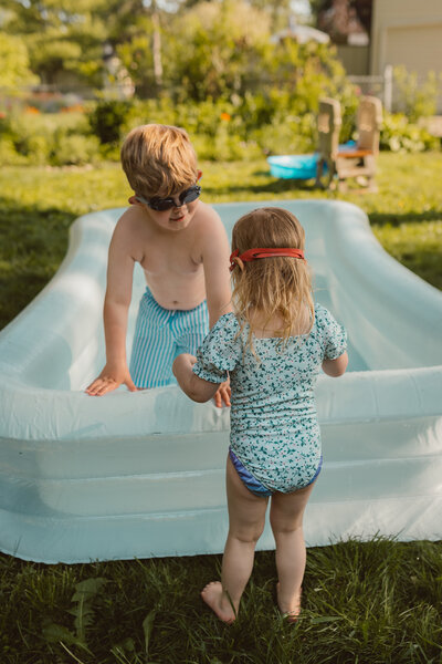 brother and sister stand at the edge of a blow up pool smiling at each other while wearing goggles