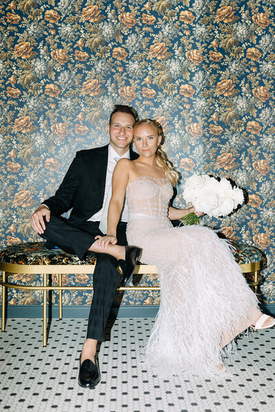 Bride and groom sit close on a bench in front of elegant floral wallpaper and pose for the camera