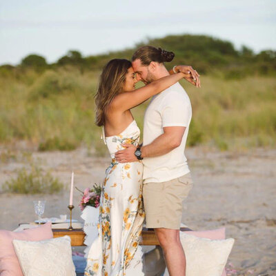 Couple embracing during a romantic beachside picnic setup with candles and pillows, styled by Yours Truly, a wedding planner and designer in New Jersey.