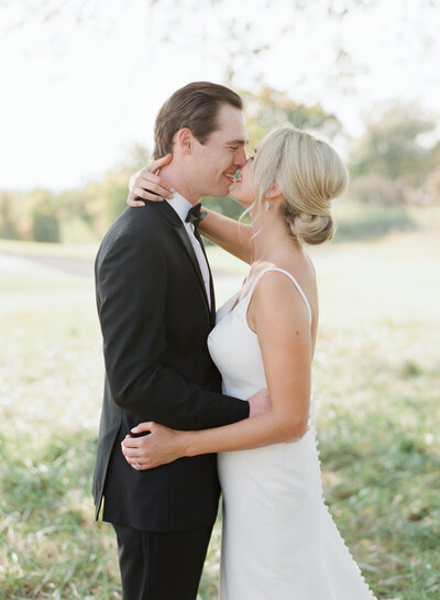 bride and groom kissing at their fall wedding under a tree with light and airy photography and the groom is wearing a black tuxedo and the bride is wearing a summer wedding dress with spaghetti straps and  a sleek back bridal up-do hairstyle with diamond earrings and photography by Tulsa Wedding Photographer Laura Eddy
