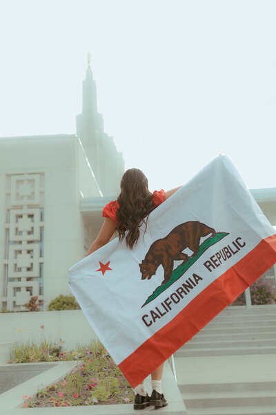 A missionary holds the California flag as she stands with her back facing the camera. She is looking at the Idaho Falls LDS temple.