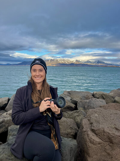 A photographer sits on a log while holding a camera and smiling off in the distance. She is surrounded by lush green grass and trees.