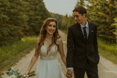 bride smiling at the camera while walking down a road holding hands with groom