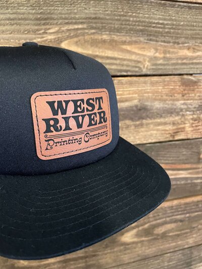 Snapback black hat with West River Printing Company logo printed on a leather patch, sewn on.