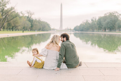 A mother and father sit on the edge of the reflecting pool in DC while their daughter peeks behind them during their photo session in Washington DC