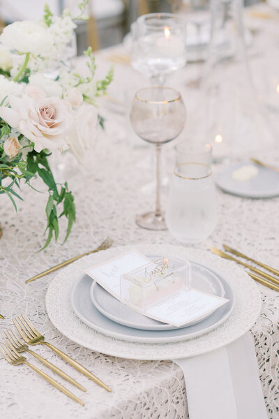 Wedding reception table setting, 3 white plates stacked together, soft pink roses, lacy table cloth, and gold cutlery.
