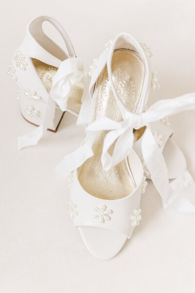 Close-up of elegant white bridal heels with floral pearl embellishments and ribbon ties, photographed on a neutral background for a Dallas-Fort Worth wedding flatlay.