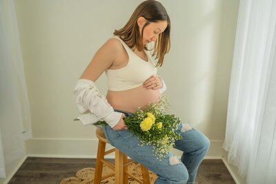 mom is white sports bra holding her pregnant belly and white and yellwo flowers while sitting on a wooden stool looking down a ther belly