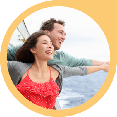 A young couple stands at the front of a ship out on open water, arms raised to either side in a classic Titanic pose. Both are laughing with pure joy.