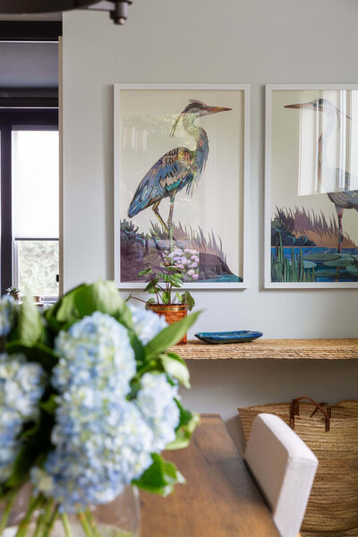 Coastal interior design featuring a framed heron artwork, a rustic woven console table, and a bouquet of blue hydrangeas in a bright, airy room.