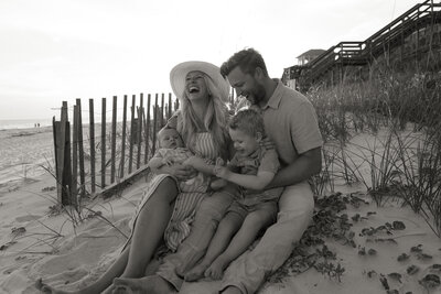 Black and white image of family of four sitting in the sand on the beach and laughing