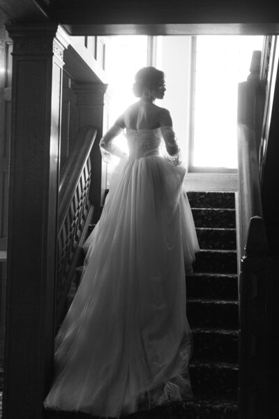 a black and white image of a bride walking of the steps of a chateau holding her dress and looking off