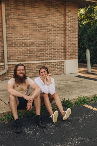 Marisa and Tanner of Msav Creative Co sit outside of a brick building on a curb and smile at the camera