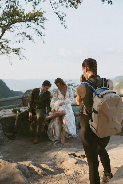 A bride and groom are sitting side by side sharing an intimate moment looking at the tattoos on the brides legs while a photographer takes photos of them.