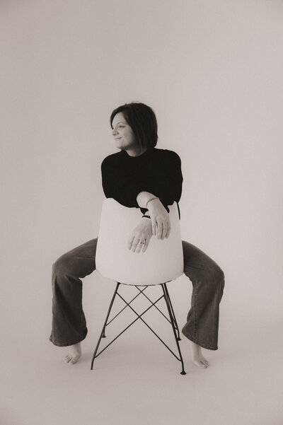 A woman sits backwards on a chair, facing right, with her arms resting on the backrest. Captured by a talented Macon family photographer, she is barefoot, wearing jeans and a black top, and looking off to the side with a slight smile.